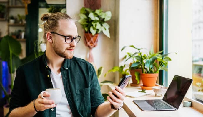 Man drinking cup of coffee using cell phone protected with Norton 360 for Mobile.